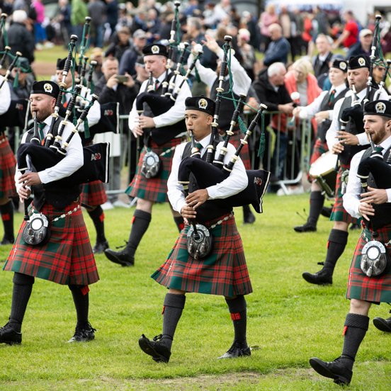 A band of pipers in red kilts march across Glasgow Green with crowds looking on