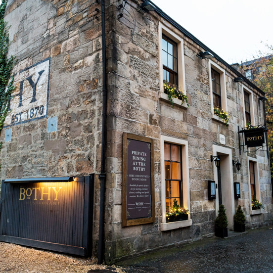 Exterior image of The Bothy restaurant shows an old, 2-storey sandstone building. The front of the building has traditional windows with dark frames, window boxes and small potted trees decorate the windows & doorway. The side of the building is half covered in ivy, obscuring a large wall painting, the "HY" of Bothy and "est.1870" are visible. A wide wooden gate, propped open against the building and a black hanging sign above the door have gold lettering reading "Bothy". The ground is paved and gravel. 