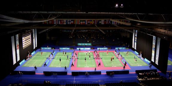 A view of five badminton courts, national flags hang from the ceiling.