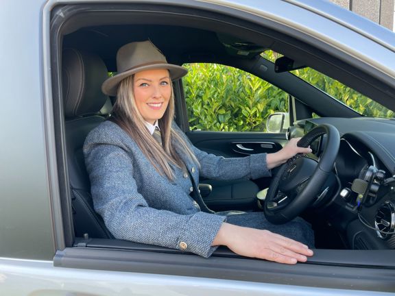 Woman sitting in driving seat of a car, window down, sliming at camera.