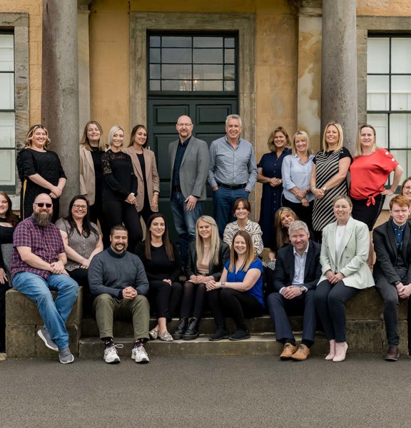 A photograph of the Fitwise team, sat and stood together on the steps of a blonde sandstone building. The steps are flagged by 2 pillars. Behind the group a large panelled door painted dark green is visible, with a small paned window above. Large, paned, sash and case windows flank the image on either side - their frames are painted in the same green. The ground is paved with tarmac, with a gravel border against the building.