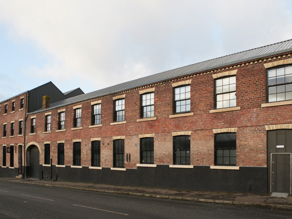 A view of The Engine Works building's exterior, lining the far side of a quiet street with narrow tarmac pavements. The building is mainly red brick with black and white brick accents. The roof is stepped with the building's left half boasting 3 storeys and the right half only 2. Large, paned, black framed windows line both halves. The roofline is framed against a grey sky.