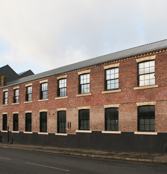 A view of The Engine Works building's exterior, lining the far side of a quiet street with narrow tarmac pavements. The building is mainly red brick with black and white brick accents. The roof is stepped with the building's left half boasting 3 storeys and the right half only 2. Large, paned, black framed windows line both halves. The roofline is framed against a grey sky.