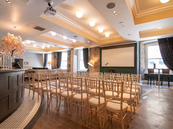 An interior view of a room in Citation. The room is laid out with 5 rows of white chairs. At the front of the room is a large projection screen and a microphone on a stand are visible. The room is bright with overhead lights, standing lamps and  4 large windows visible; dark heavy curtains frame them. On the left, the edge of a curving wooden bar is visible. Beer taps, a till and a branch covered in blossom are all visible on top of it.