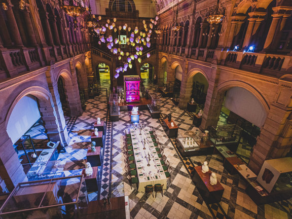 An internal view of the Kelvingrove East Court, set up for a private dinner, taken from a first storey balcony. A long, vaulted, room adorned with carved sandstone walls and a polished-stone floor. An artwork of white heads is suspended from the ceiling, the event's lighting makes them look pink and yellow. Down the centre of the floor is a long table covered in an ivory tablecloth, wooden chairs surround it, the table is set and decorated. Glass vitrines, carved busts and other objects surround the table.