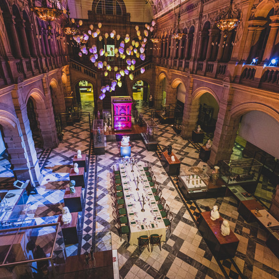 An internal view of the Kelvingrove East Court, set up for a private dinner, taken from a first storey balcony. A long, vaulted, room adorned with carved sandstone walls and a polished-stone floor. An artwork of white heads is suspended from the ceiling, the event's lighting makes them look pink and yellow. Down the centre of the floor is a long table covered in an ivory tablecloth, wooden chairs surround it, the table is set and decorated. Glass vitrines, carved busts and other objects surround the table.