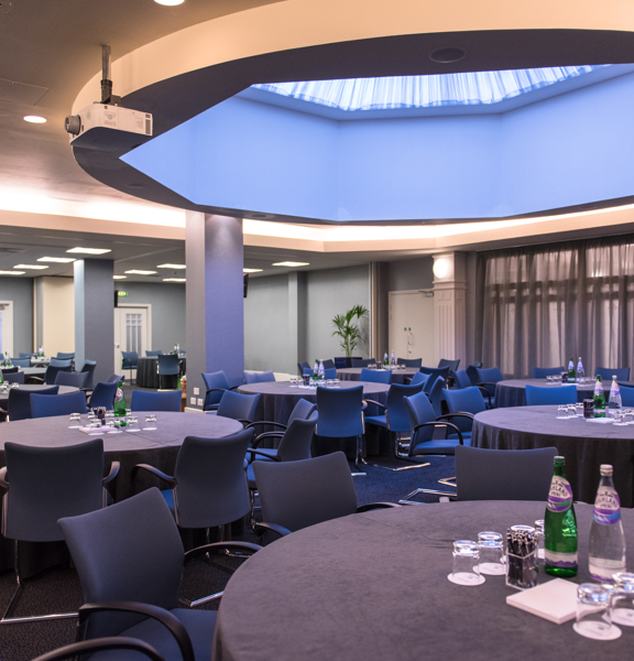 An interior image of a large room at the Golden Jubilee hotel. The room is predominantly decorated in shades of grey, and the windows on the left hand wall are covered with translucent grey curtains. Ceiling panel lights and an octagonal skylight light the room. Grey and blue fabric chairs are arranged around circular tables. They have grey table cloths, bottled water, glasses and stationary. 2 projectors can be seen in the ceiling, pointing out of shot. 3 doorways can be seen out of the room.