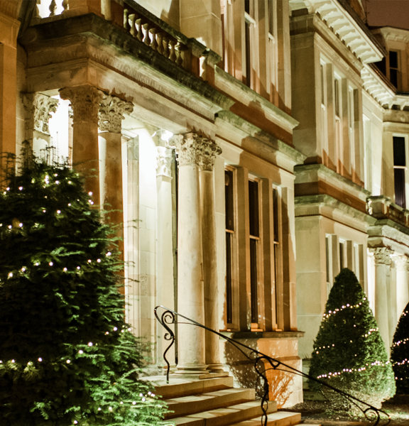 An external view of Hotel Du Vin shows a grand terrace of Victorian sandstone town houses. It is night and the building is up-lit showing the buildings carved Roman Tuscan pillars and lintels. Small box-hedges trees cut into cones line the length of the terrace's pavement and are decorated with a spiral of fairylights. An ornate, black handrail can be seen on the stairs to the entrance. The doorway is lit and though the blind is half down, a view through the front window reveals a lit, wooden panelled room.