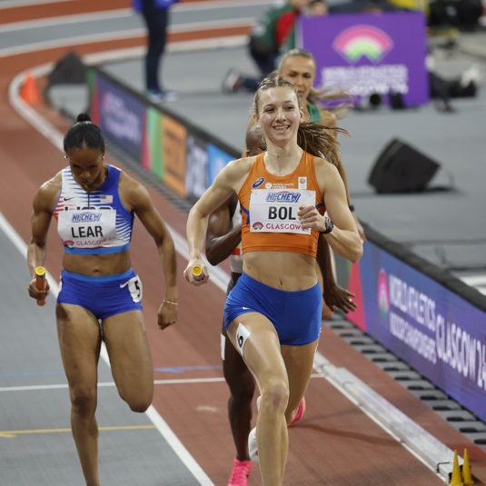 An athlete wearing an orange top runs on track, smiling.