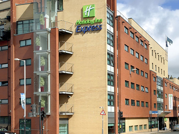 An exterior image of the Holiday Inn Express Riverside shows a modern yellow and red brick multi-storey building, from the viewpoint it is possible to see the building is on on the corner of 2 roads. A large sign on the end of the building reads "Holiday Inn Express" in the brand's green and blue. An entrance can be seen down the right-hand road, with a white sign marking it. The building is seen against a blue cloudy sky.