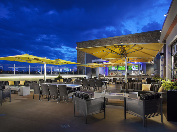 An image of the TopGolf Terrace Bar during the evening. A large paved balcony area is filled with cushioned, metal sofas, woven high-backed chairs, polished stone tables and large, square, yellow parasols. The light-brick building attached, has 2 huge cut away walls to the interior. A stone-effect bar, large TV screens and further seating can all be seen within. 