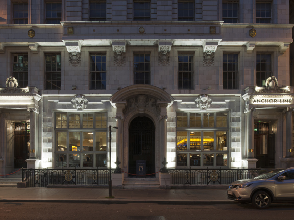 An exterior photo of The Anchor Line taken at night from across the road, depicts the first 3 floors of a grand, white Victorian building with large traditional windows.  The building is adorned with carved and gilded details & boasts 3 large carved doorways. The right hand one is lit and has dark lettering that reads "Anchorline" on its lintel. Through the window a bar can be seen lit up. The pavement outside is paved, street signs, lampposts and a parked car can be seen outside.