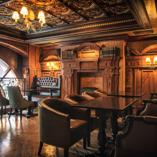 An interior image of the ornately decorated "Library" space. The room has polished wooden floors and intricately carved wall and ceiling panels. Brass ceiling lights and wall sconces with small lampshades light the space with golden light. The far, left wall is dominated by the top arch of a large window. Dark wooden tables and traditional, upholstered chairs and benches line the edge of the room, besides a large, ornate fireplace flanked by leather bound books.