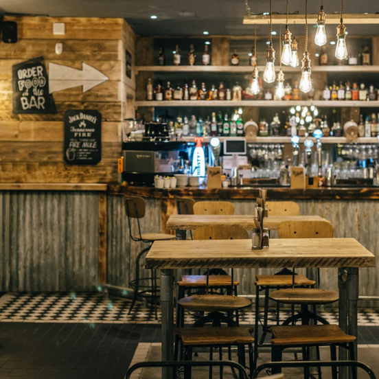 An image from inside the Van Winkle restaurant and bar, shows a room with rustic, reclaimed wood & corrugated metal panelling on the walls. The floor is tiles in a mix of black and white. High wood and metal stools sit at high scaffolding-style tables and counters. Bare, filament lightbulbs with brass fittings hang in groupings. A well stocked bar and shelves takes up much of the back wall, and small wall mounted blackboard signs list offers and direction.