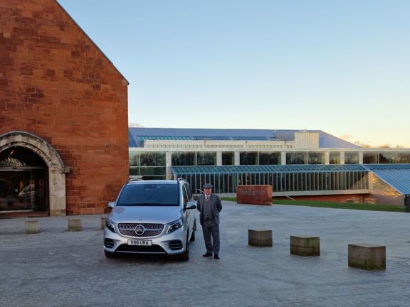 Silver Mercedes people carrier parked in front on museum called Burrell Collection. Chauffeur driver standing next to car. 