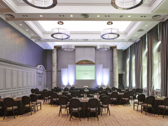 A photo of the voco Grand Central ballroom shows a traditional, high-ceilinged room, with a beige chevron patterned carpet throughout & walls have white painted wooden panelling. 5 large, curtained windows line the right wall. The far wall boasts 2 large marble pillars either side of a large projection screen. Large round tables with brown cloths, and wooden chairs with matching leather seats are arranged into groups. The ceiling has traditional plaster moulding contrasted with modern, glass light fixtures,