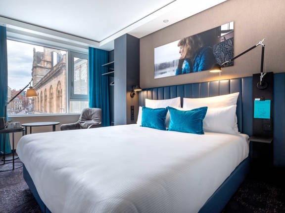 An interior from a Motel One bedroom. A bright hotel room with dark grey patterned carpet and a large, bed with white sheets and a teal leather headboard. A large window takes up a majority of the far wall, a facade of a large sandstone building can be seen outside. A coffee table and a grey armchair are positioned in the window on the far side of the bed. A large photographic print above the bed depicts a young, white woman gazing out of a window. Lamps, mugs, hangers and plug sockets are also visible.