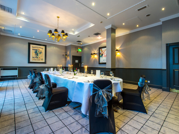 An interior shot of a meeting space at Holiday Inn Theatreland shows a room with taupe walls and dark panelling, the floor is carpeted with a gridded pattern on it. The centre of the room has a long table with a white table cloth; wine bottle, candle sticks and glasses are all visible. Several black-covered chairs surround the table, they are decorated with organza bows. 2 windows can just be seen on the most left-hand wall, sconces, a ceiling light and spotlights also help light the room. 