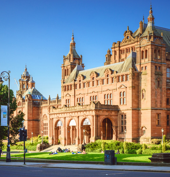 An exterior view of part of the Kelvingrove museum on a sunny day, shows the ornate, Victorian red-sandstone facade of the main entrance. The building is heavilyadorned, the main entrance is dominated by 3 large archways at the top of a few steps. The roof line is decorated with pinnacles, buttresses and turrets. In front of the museum, a sumptuous green lawn, shrubs and trees line the pavement and road in the foreground of the image. A pedestrian crossing and an ornate lamppost are also visible.