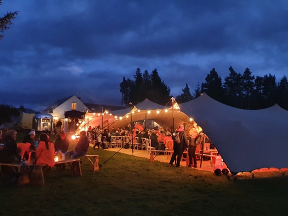 Large glamping stretch tent with festoon lights hanging from the ceiling. Group of People inside seated at dinning table and people outside sitting on benches around a campfire at night.