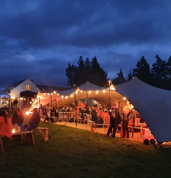 Large glamping stretch tent with festoon lights hanging from the ceiling. Group of People inside seated at dinning table and people outside sitting on benches around a campfire at night.