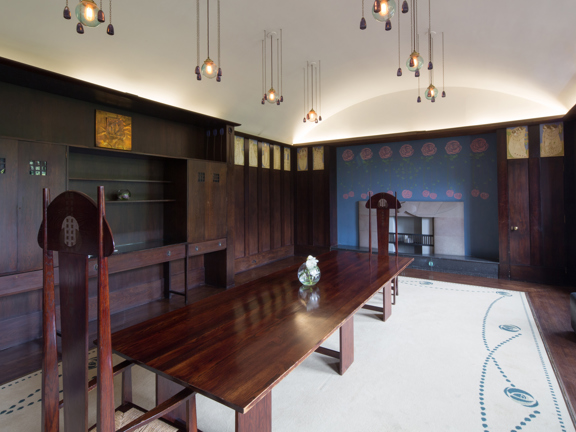 An internal view of a small dining room at House for An Art Lover. A narrow room with dark wood panelling, shelves and flooring. A cream rug with a graphic, dotted border almost fills the floor. The far wall is painted a deep blue with a repeating Glasgow-Style geometric floral motif. There is a long, bare dark wood table and 2 exceptionally tall, sculptural chairs in the same wood. The ceiling is vaulted and painted white. The ceiling lights, painted wall panels and a brassy moulding add further detail.