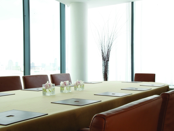 An interior view from one of Hilton Garden Inn's meeting room shows the corner of a bright room with a white ceiling, it has floor to ceiling windows on both visible walls and a cyclindrical, white pillar in the corner. A long table, covered with a light-ochre tablecloth runs through the image, surrounded by tan leather chairs. Leather placemats and modern flower arrangements decorate the space. A view of the city can be seen out of the left window, while the right one is covered in a white mesh curtain.
