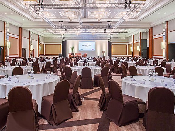 An interior view of the Crowne Plaza ballroom shows a large room decorated in neutral tones of brown and beige. The floor is filled with large round tables, covered with white table cloths and chairs around them with brown fabric covers. At the far end of the room a stage with 3 seats and a podium is evident, behind which a projection screen is pulled down. There are chandeliers in the ceiling as well as a technical light rig. 