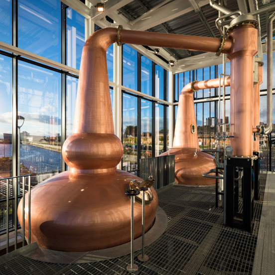 A view from inside the Clydeside Distillery. A room with glass walls, it is taken in daylight from a first-storey mezzanine. The space is dominated by 2 enormous, copper whisky stills - an important instrument in the distilling of whisky. They have bulbous forms and highly polished surfaces, copper and silver pipes lead off of them. A black railing runs the length of the mezzanine, the floor is a mix of metal grates and tread plate. Out of the windows views of the River Clyde and blue skies are visible. 