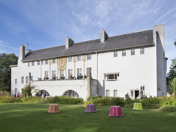 An external view of Glasgow's House for An Art Lover, a Charles Rennie Mackintosh building with 3 storeys, white rendering, blonde sandstone art-nouveau carved details and a shallow, dark peaked roof. A large balcony stretches out of the first floor into a manicured garden, there is a lawn and a wide flower bed between the viewer and the building. People sit on the terrace and mill around a french window entrance in the ground floor. Jelly mould shaped sculptures in bright colours, dot the lawn.