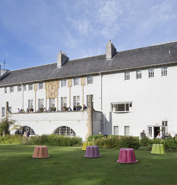 An external view of Glasgow's House for An Art Lover, a Charles Rennie Mackintosh building with 3 storeys, white rendering, blonde sandstone art-nouveau carved details and a shallow, dark peaked roof. A large balcony stretches out of the first floor into a manicured garden, there is a lawn and a wide flower bed between the viewer and the building. People sit on the terrace and mill around a french window entrance in the ground floor. Jelly mould shaped sculptures in bright colours, dot the lawn.