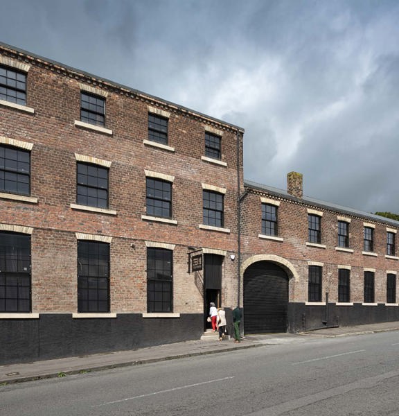 A view of The Engine Works building's exterior, lining the far side of a quiet street with narrow tarmac pavements. 3 people are climbing the small stone steps into the main entrance. To their right is a large arched entrance way with a black shutter. The building is mainly red brick with black and white brick accents. The roof is stepped with the building's left half boasting 3 storeys and the right half only 2. Large, paned, black framed windows line both halves. The roofline is framed against a grey sky.