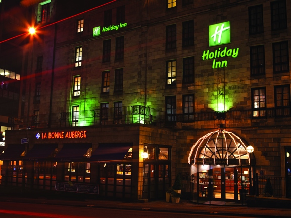 An exterior view of Holiday Inn Theatreland by night shows a 6 storeyed stone building. The building is uplit on the first floor with green lights and 2 lit signs on the facade read "Holiday Inn" and bear the companies logo in green. The main entrance is a glass, sliding door, with an arched window above. On the ground floor a space with large, windows and awnings juts out from the rest of the building; it has red neon signage reading, "La Bonne Auberge." To the left streetlights and buildings are visible.