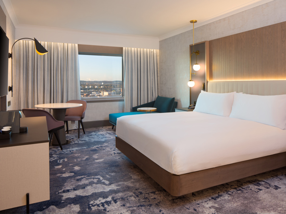 An interior view of an Executive King room at Glasgow Hilton shows a room with a mottled-grey, patterned carpet. The large bed is against the right wall and is made with crisp, white sheets, the headboard is cream and padded. In the far wall a window frames a view of the city and hills beyond, cream curtains are pulled back on either side. A small table with 2 chairs and a teal chaise chaise longue fill the space between the bed and the window. A large television is visible on the left wall over a dresser.