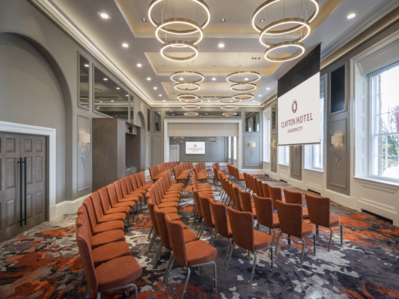 An interior view of a room in the Clayton Hotel. The room is arranged with orange chairs in 5 long rows. The room is decorated with patterned grey and orange carpets and modern circular light fittings, contrasting with original wooden panelling, sash windows and decorative plasterwork. A large television is visible on the far wall, while a projection screen hangs in the centre of the room, both are showing the Clayton Hotel branding. A double door into the room is visible on the left-hand side.