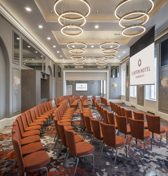 An interior view of a room in the Clayton Hotel. The room is arranged with orange chairs in 5 long rows. The room is decorated with patterned grey and orange carpets and modern circular light fittings, contrasting with original wooden panelling, sash windows and decorative plasterwork. A large television is visible on the far wall, while a projection screen hangs in the centre of the room, both are showing the Clayton Hotel branding. A double door into the room is visible on the left-hand side.