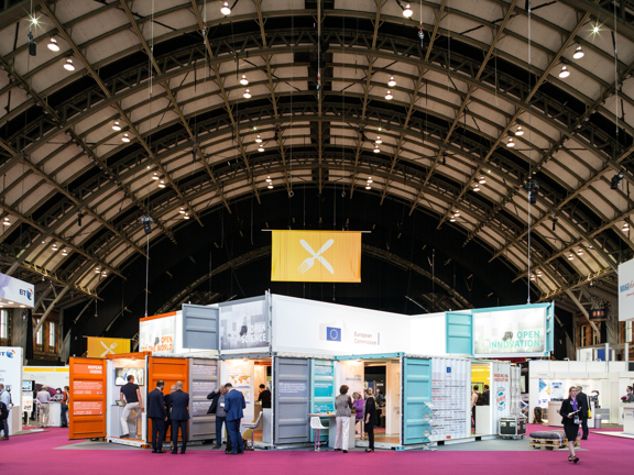 An image of the interior of a large event space with a conference build in-situ. The stalls are made up of repurposed shipping containers painted in bright colours and decorated with banners, text, boards and furniture. The space is cavernous, like a hanger with an arched roof. Technical lighting rigs and structural awnings criss-cross the curved roof - a large banner hangs form one. A pink corridor of carpet runs between the stalls.