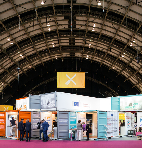 An image of the interior of a large event space with a conference build in-situ. The stalls are made up of repurposed shipping containers painted in bright colours and decorated with banners, text, boards and furniture. The space is cavernous, like a hanger with an arched roof. Technical lighting rigs and structural awnings criss-cross the curved roof - a large banner hangs form one. A pink corridor of carpet runs between the stalls.