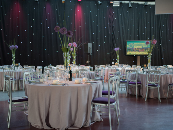 An interior image of the Glasgow Science Centre decorated for an event. The walls are covered in black fabric. Round tables are covered with cream table cloths, they are surrounded by silver chairs with plush purple seats. The tables have tall vases of alliums at their centres, with candles, glasses and silverware also decorating them. A technical lighting rig can be seen at the top of the far wall, a large television screen on a stand and the bottom corner of a projection screen are also visible. 