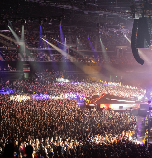 A huge audience in the OVO Hydro watching a band on the stage with blue and  white lights shining on parts of the audience