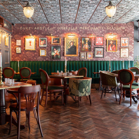 The richly decorated dining room of the Duke's Umbrella. The restaurant is darkly decorated with dark-wood parquet flooring and panelling. Deep red botanical wall paper decorates the far wall, with gilt framed prints and brass spotlights curated on it. A high-backed deep-green upholstered bench lines the far wall. The room is otherwise furnished with dark wood tables and eclectic chairs. Plate glass windows allow daylight to stream in, while cut-glass light features are mounted to the carved-wood ceiling.