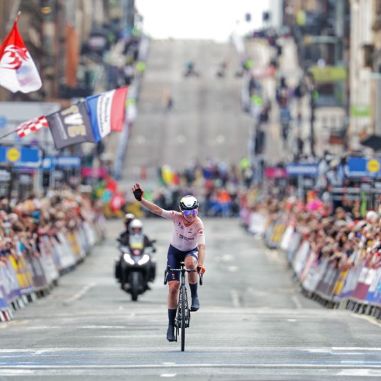 A female cyclist on the road near the finish line waving as crowds are to either side of her waving various countries flags at the UCI Cycling World Championships