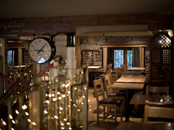 An interior view of the Stravaigin restaurant's mezzanine level. The space has a low, beamed ceiling, supported by glossy black and white pillars. A traditional white-faced clock is mounted to one the beams at the top of the stairs. The railings around the mezzanine level are decorated with white fairy lights. Wooden tables and chairs are visible around the edge of the space. The floor is wooden, the far wall is bare stone and has a small paned window.