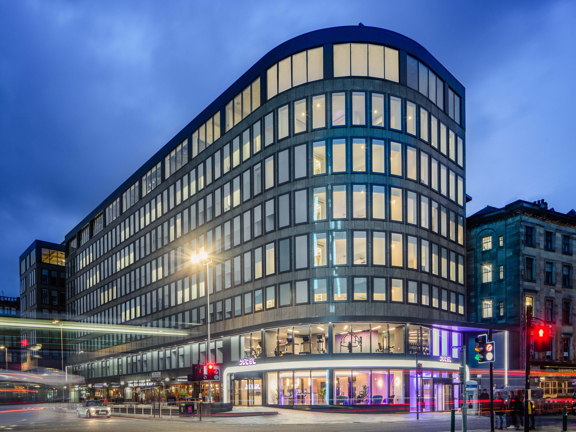 An external photo of the curved glass facade of the Yotel hotel. The hotel sits on the far corner of a wide road junction at dusk, the photographer has used a long exposure and vehicle lights have left streaks of light across the image. The hotel is an 8 floored grey building, covered in long, lit rectangular windows with dark frames. The pavement outside the hotels entrance is sloped and paved. An older sandstone buildings sits adjacent to it on the right and a modern building to the right.