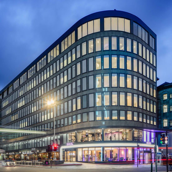 An external photo of the curved glass facade of the Yotel hotel. The hotel sits on the far corner of a wide road junction at dusk, the photographer has used a long exposure and vehicle lights have left streaks of light across the image. The hotel is an 8 floored grey building, covered in long, lit rectangular windows with dark frames. The pavement outside the hotels entrance is sloped and paved. An older sandstone buildings sits adjacent to it on the right and a modern building to the right.