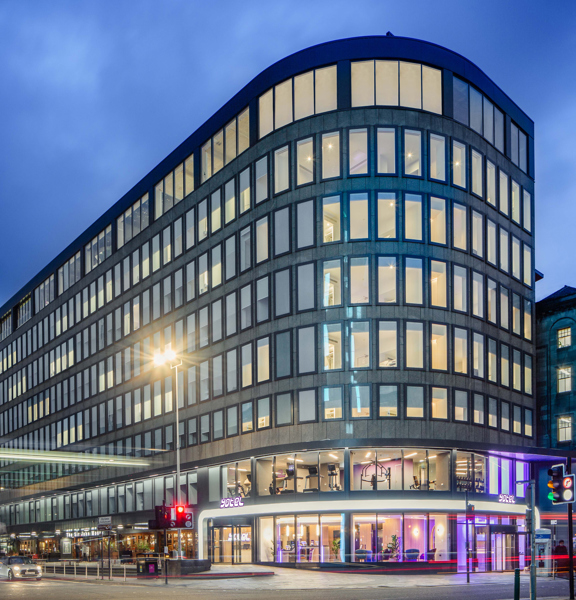 An external photo of the curved glass facade of the Yotel hotel. The hotel sits on the far corner of a wide road junction at dusk, the photographer has used a long exposure and vehicle lights have left streaks of light across the image. The hotel is an 8 floored grey building, covered in long, lit rectangular windows with dark frames. The pavement outside the hotels entrance is sloped and paved. An older sandstone buildings sits adjacent to it on the right and a modern building to the right.