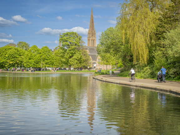 Sunny view of a small, tree-lined pond in Queen's Park, with cyclists and people strolling along a path, with a church spire in the background