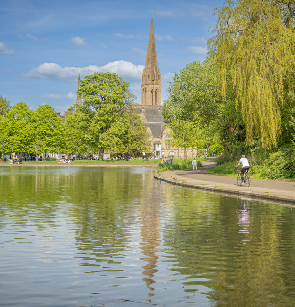 Sunny view of a small, tree-lined pond in Queen's Park, with cyclists and people strolling along a path, with a church spire in the background