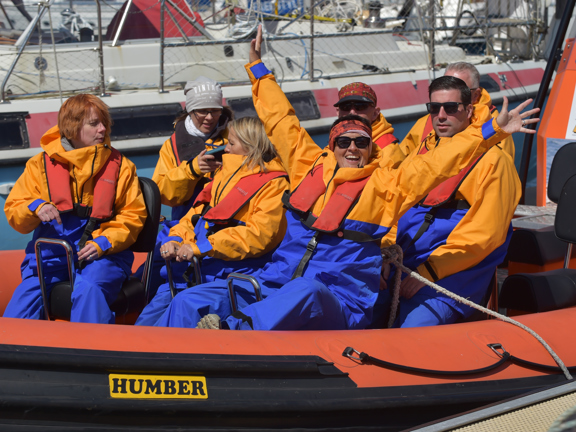 Group of seven people on an orange and blue speedboat wearing matching orange and blue waterproof outfits. Front person is waving and smiling at camera.