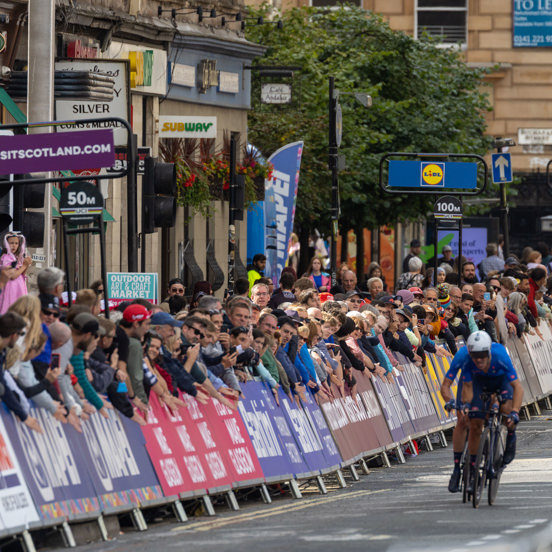 Cyclist in the road race wearing a blue jersey going past a large crowd to the left of the photograph